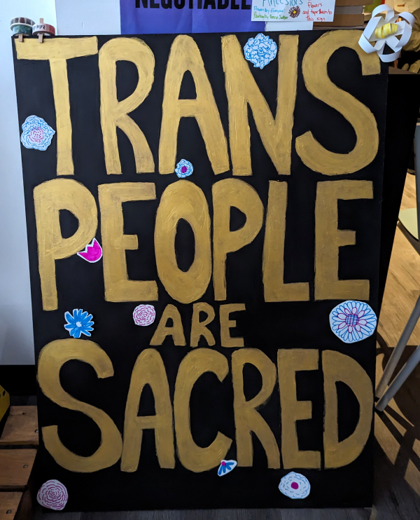 Trans People Are Sacred - a hand lettered sign with gold letters on a black background, decorated with handmade flowers